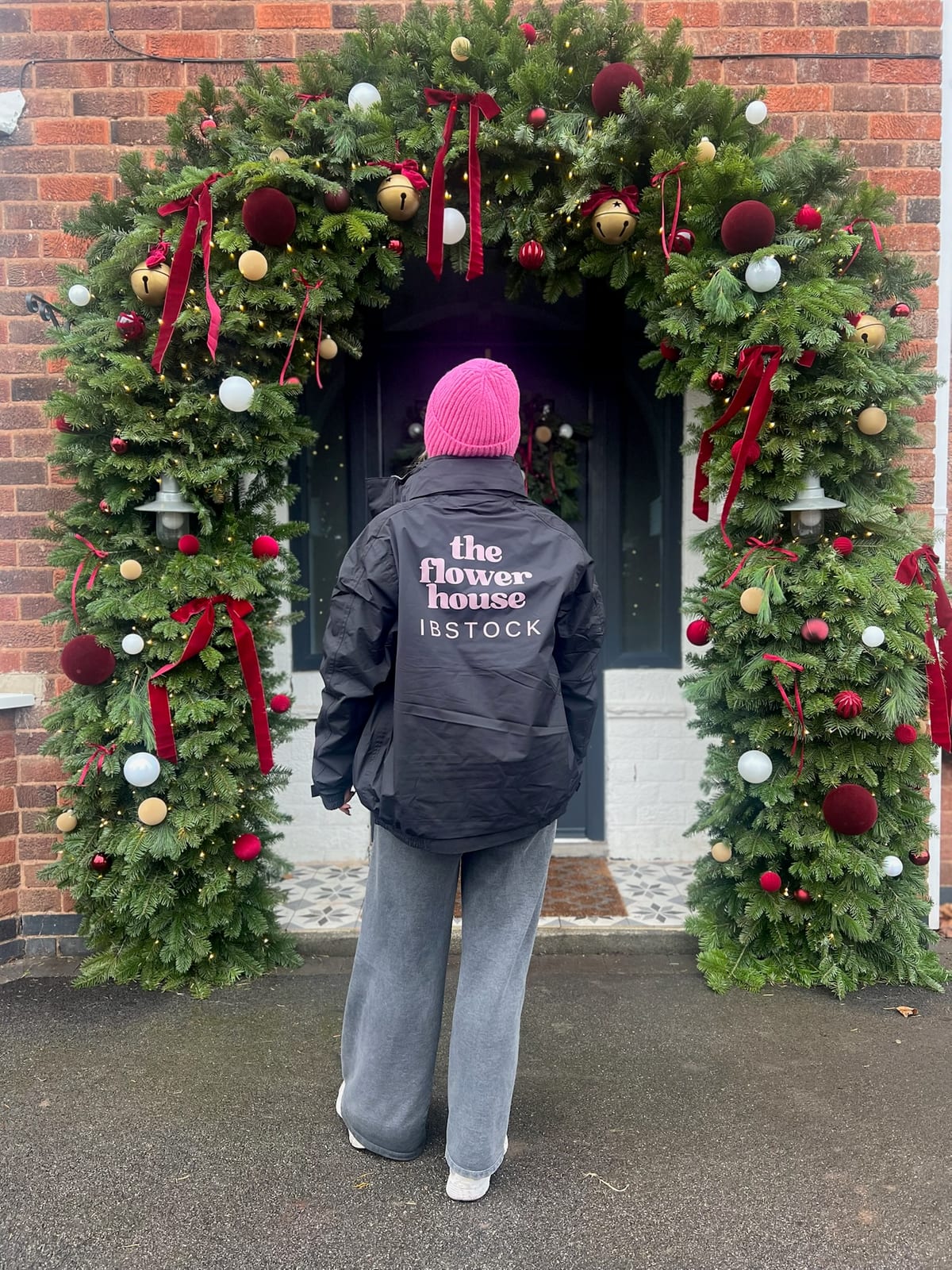 A florist in a branded jacket in front of a brick doorway framed by a garland arch with red velvet ribbons, brass bells and burgundy, white and gold baubles.
