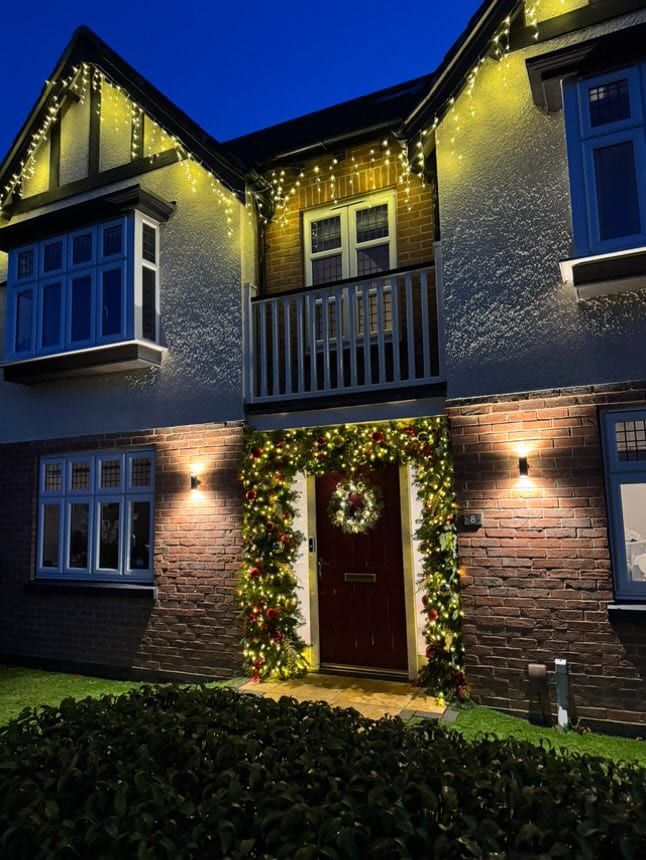 House exterior at night with lit garland framing the front door and icicle lights along the roofline.