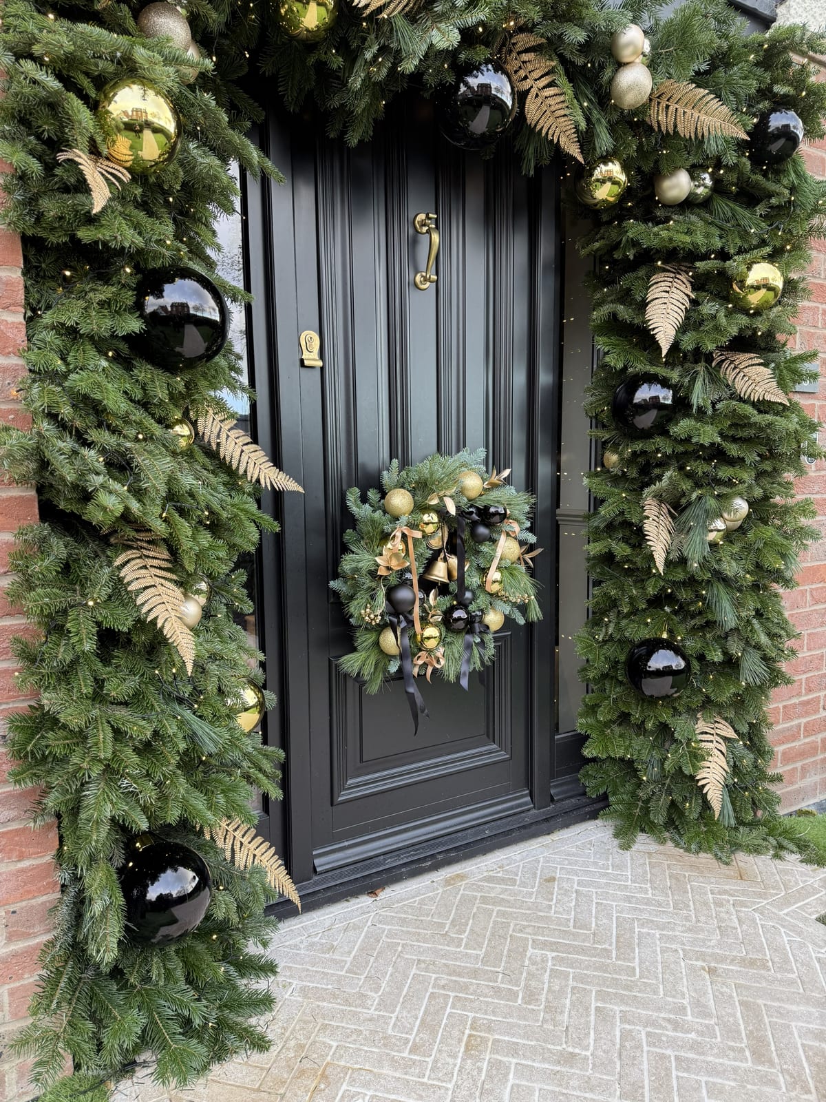 Black front door framed by a garland arch with gold ferns, gold and black baubles, and a matching wreath.