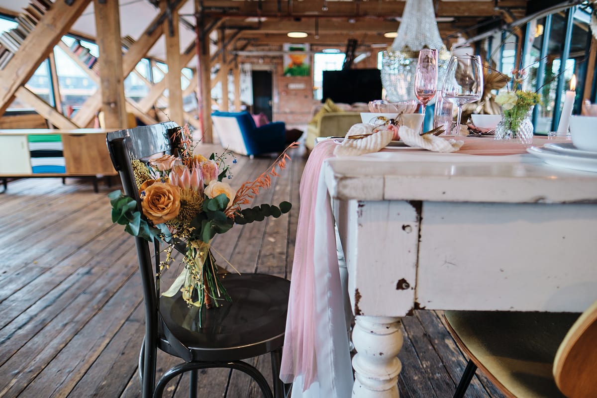 Long banquet table in a beamed barn, dressed with peach and cream arrangements and trailing blush ribbon.