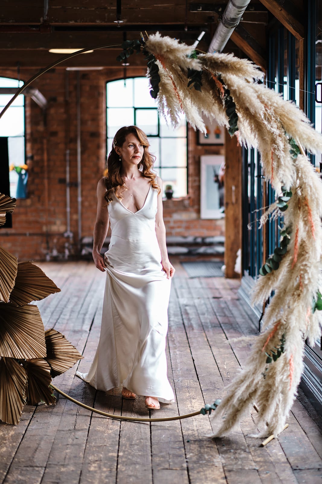 Bride in a slip dress standing beside a tall arch of dried grasses and foliage in a converted barn.