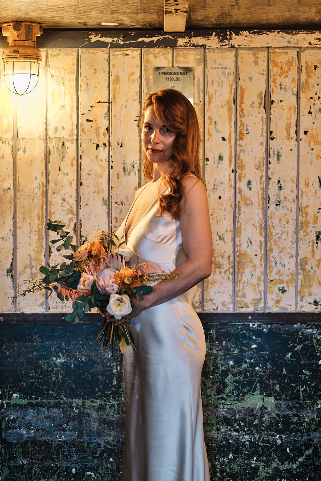 Bride holding a loose bouquet of peach roses and foliage in front of weathered wooden doors.