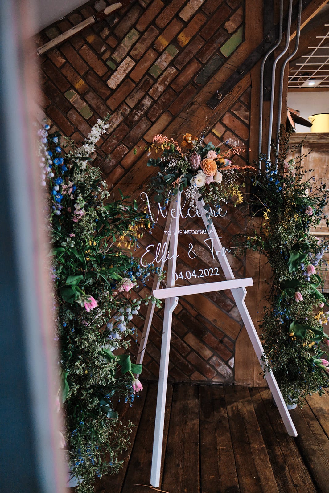 Wooden easel framed by an arrangement of foliage and seasonal flowers against an exposed brick wall.