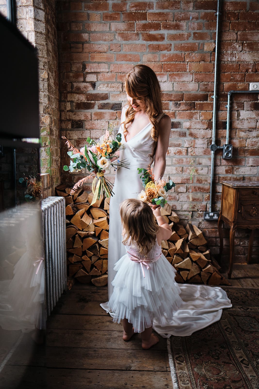 Bride and a flower girl beside an exposed brick wall, both carrying peach and cream flowers.