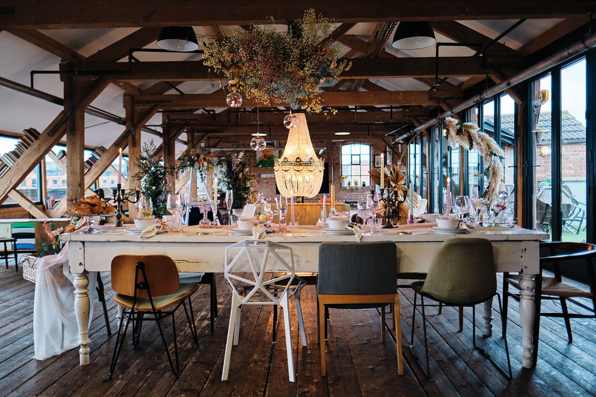 Beamed barn set for a wedding breakfast, with a long dressed table and hanging chandelier.