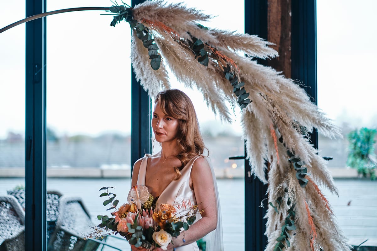 Bride beside a tall arch of dried grasses in front of a window in a converted barn.