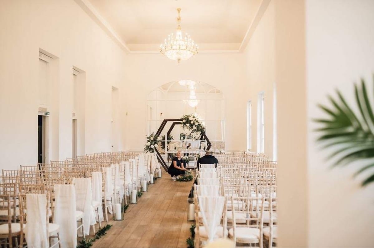 Ceremony room with rows of chairs facing a dark hexagonal arch dressed in white flowers and foliage.