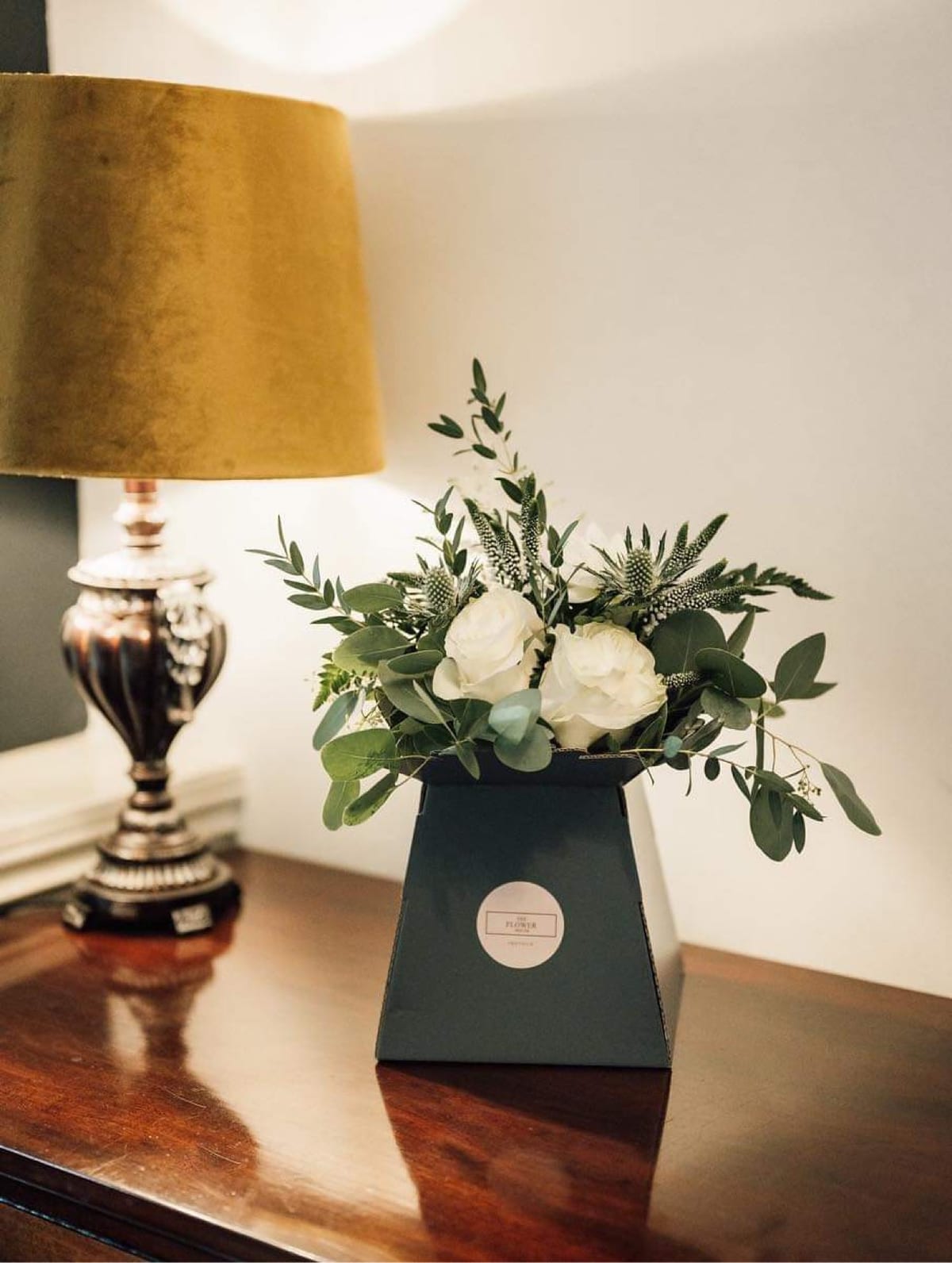 Black vase of white roses and eucalyptus on a side table beneath a gold lamp.