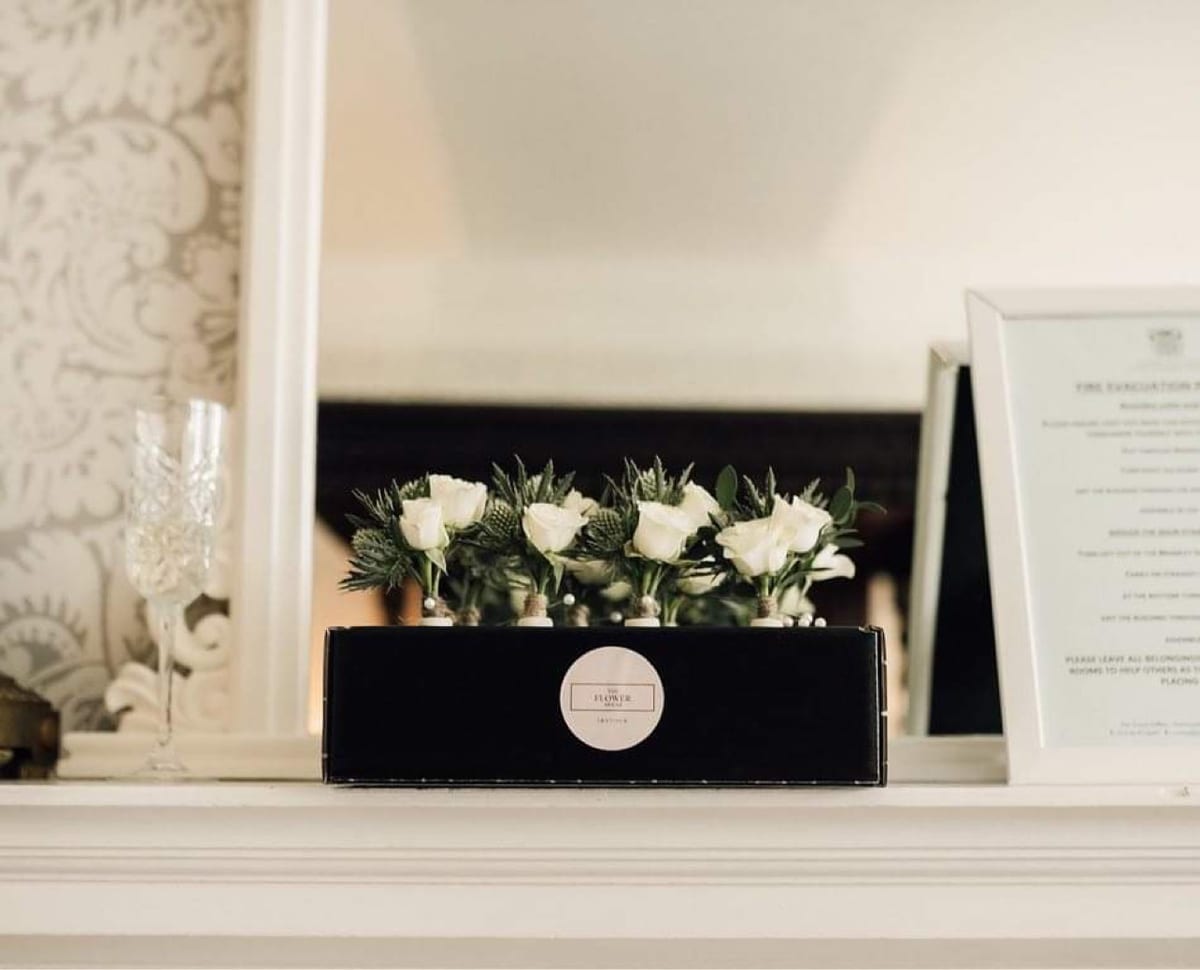 Black planter of white roses and eucalyptus on a mantelpiece in front of a mirror.