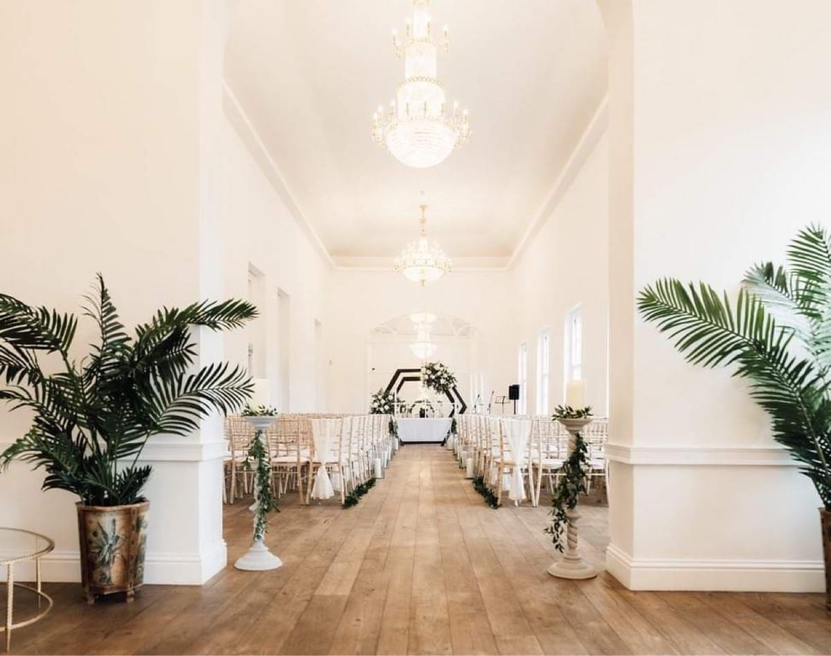 Light ceremony room with tall urns of palm leaves and greenery flanking the aisle.