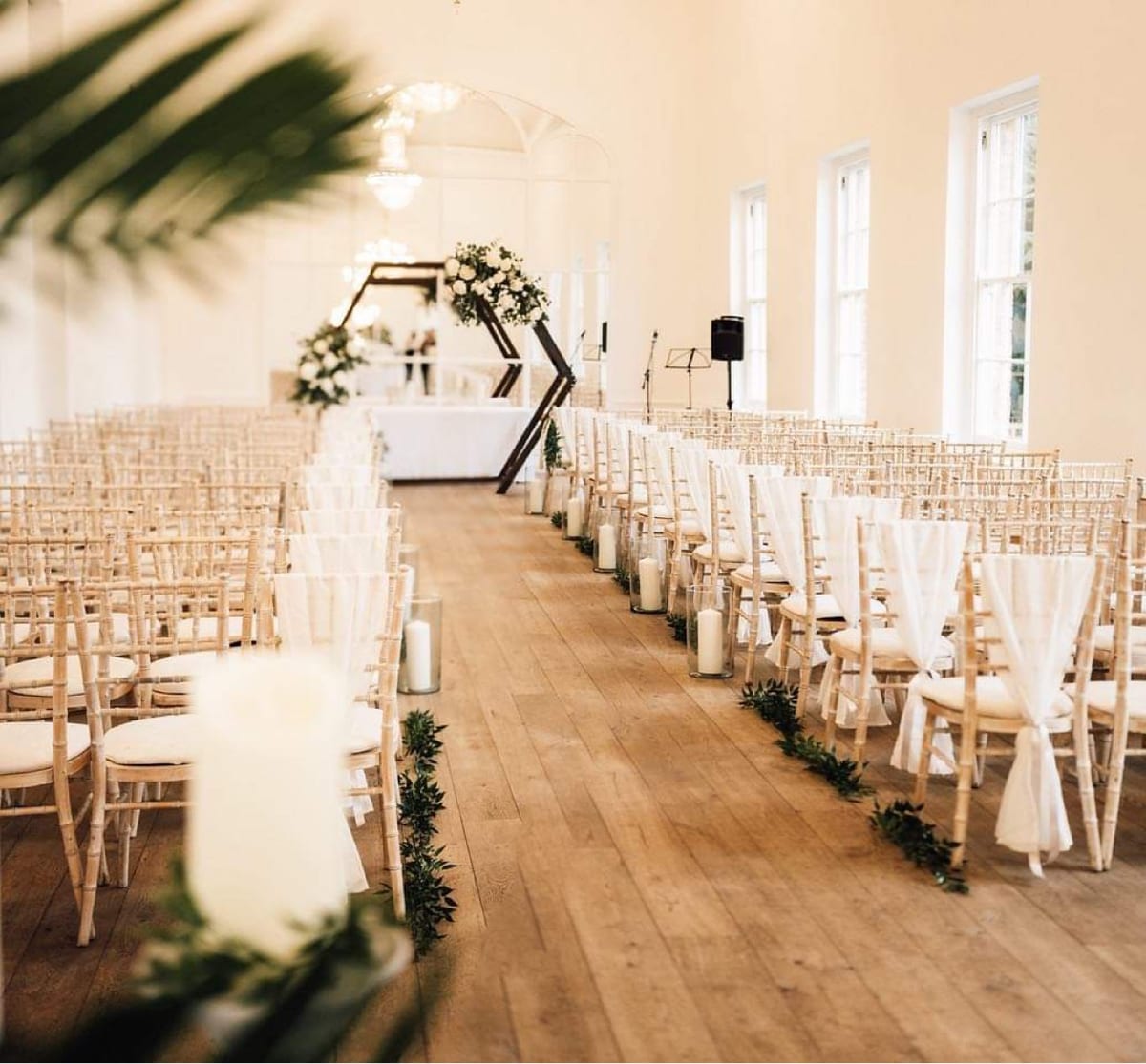 Ceremony aisle lined with chairs, greenery and a pillar candle on a wooden floor.