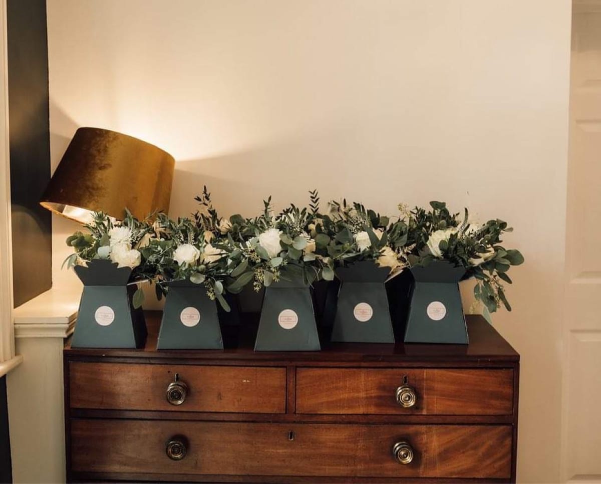 Black boxes of white roses and foliage arranged along an antique chest of drawers.