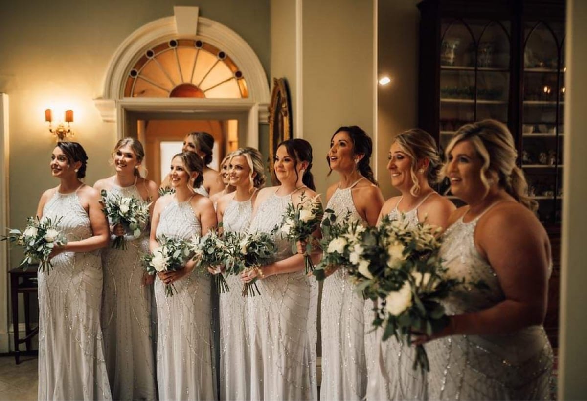 Bridesmaids in pale dresses holding bouquets of white roses and foliage.