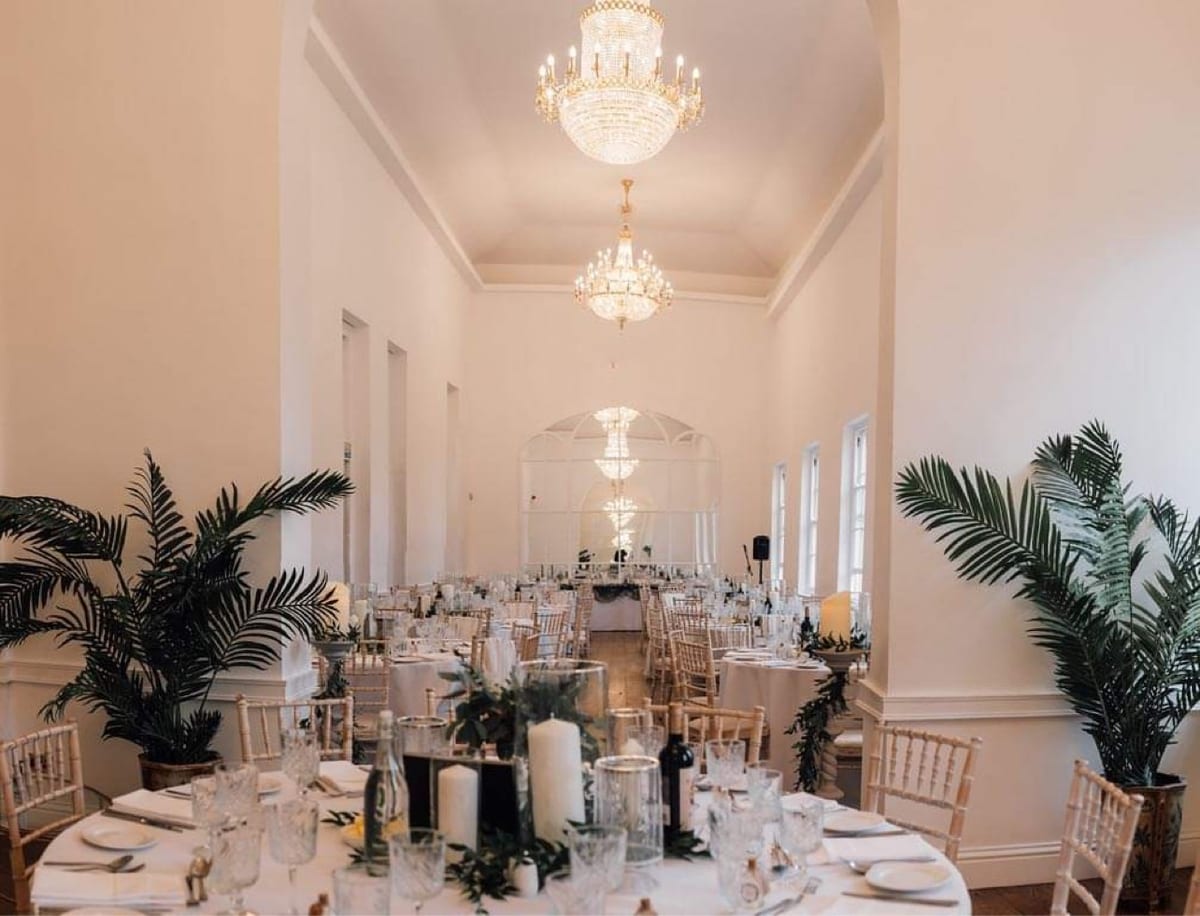 Reception room set for dinner, with tall greenery and chandeliers overhead.