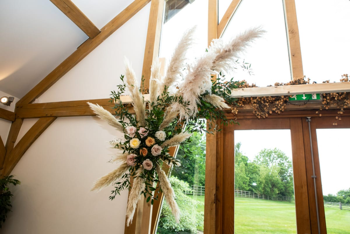 Corner of a ceremony arch dressed with dried grasses, peach roses and foliage against oak beams.