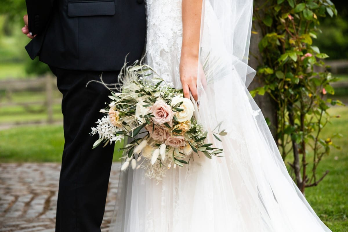 Bride holding a bouquet of peach and cream roses with dried grasses, standing beside the groom.