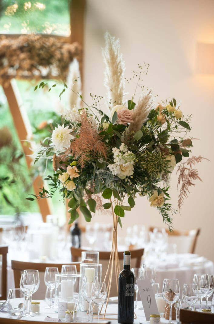 Tall centrepiece of peach roses, dried grasses and foliage on a set dining table.