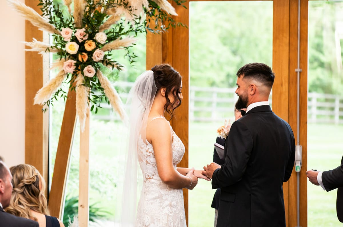 Couple facing each other at the ceremony beneath an arch of dried grasses and flowers.