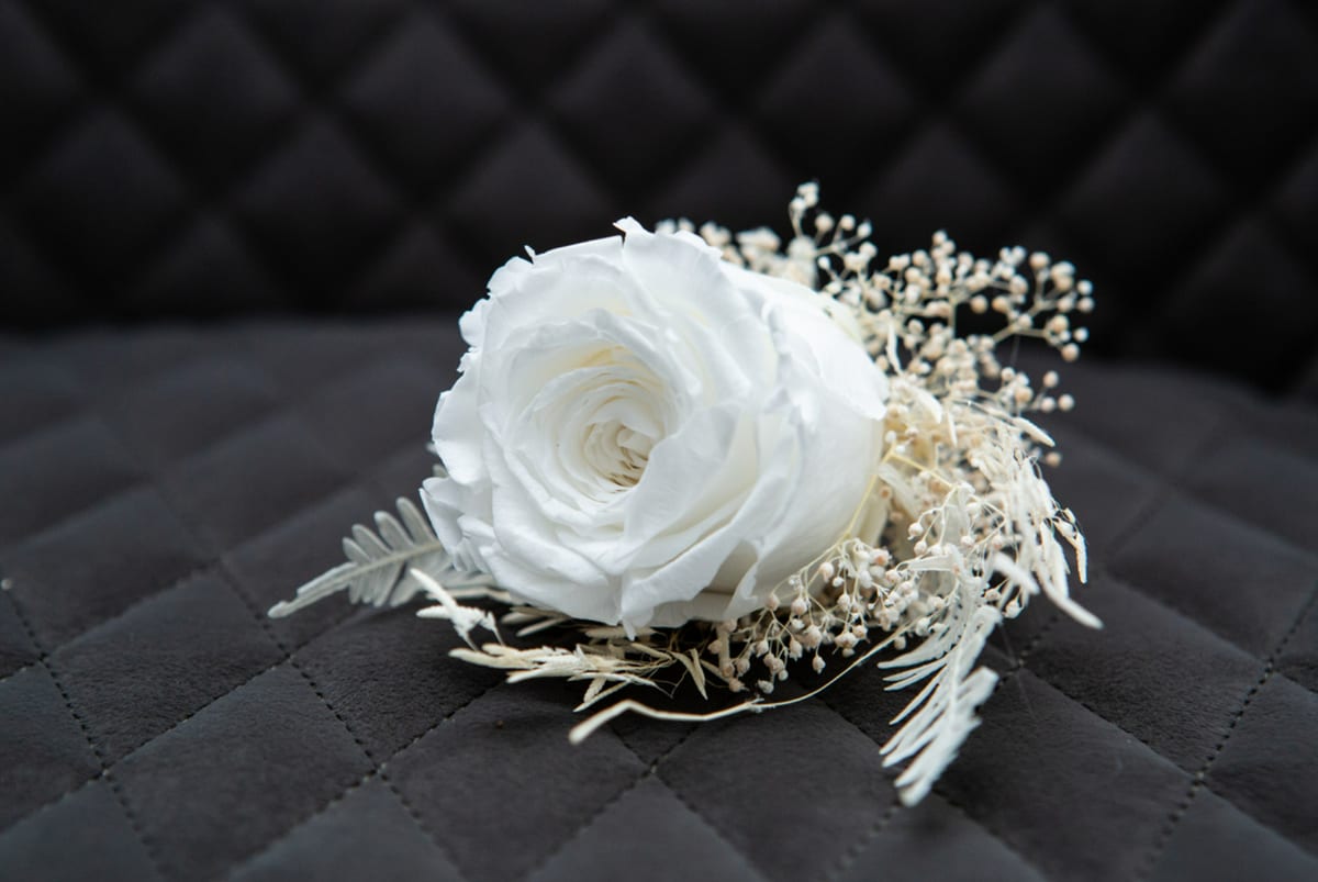 Buttonhole of a white rose with dried grasses and fine white filler flowers.